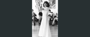 Woman dark shoulder length hair wearing a long white dress claps hands high as she dances for audience. Black and white photograph.