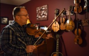 Bob Cathey wears thin framed glasses and a blue and white plaid button down shirt. Bob is tuning a violin and behind him are a shelf of other violins.