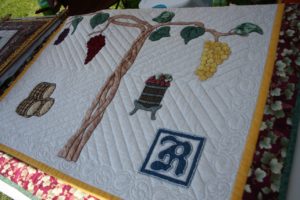 Lois Mueller's vineyard quilt. The quilt is a white background with a red leaf border. It features a design of grape branches, one side red and the other yellow. Alongside the grapes are barrels and the Robller Vibeyard logo.