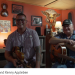 John P. Williams and Jenny Applebee pose for photo during an interview in Williams' home. Both men wear thin wired glasses with plaid button down shirts and denim jeans. WIlliams balances a fiddle in his lap wwhile Applebee balanaces a guitar.