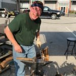 Mike McLaughlin wears thin framed glasses, a dark green crew neck shirt and denim jeans. He wears a hat as well which is dark blue. Mike is facing the camera with a smile on his face as he works on a blacksmithing project.