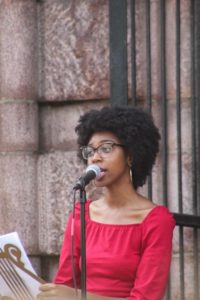 Photographed is Brianna Chandler, a young Black woman. She has combed out afro hair, a pair of gold hoop earrings. Brianna wears black framed glasses and a red longsleeve top that shows her collar bones. She is speaking into a microphone.