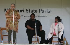 Photographed during a live performance, master storytellers: Loretta Washington, Angela J. Williams, and Gladys Caines Coggswell. Standing at the microphone is Loretta Washington, an elderly light-skinned Black woman with short cut salt and pepper hair. She wears a long earth toned patterned shirt and green pants, brown sandals. To her left sits Angela J. Williams, a plus sized darker-skinned Black woman wearing a quarter sleeved black shirt and long black pants. Beisde Angela sits Gladys Caines Coggswell, a middle age Black woman with shoulder length straight hair. She wears a pink shirt tucked under a white cardigan, black pants, and black shoes. All three woman are behind microphones.