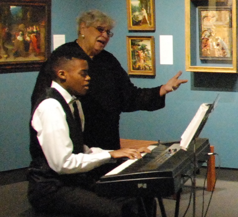 Master gospel vocalist and pianist Doris Frazier photographed performing with her apprentice Peyton Boyd in 2016. Doris Frazier stands behind her apprentice wearing a long sleeve all black outfit, she is an elderly lightskinned Black woman with short cropped grey hair. Her apprentice Peyton Boyd appears beside her wearing a tuxedo top and black bottoms, he is a younger Black male. Peyton Boyd is playing the electric keyboard and singing.