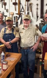 Photographed in studio, James Price and Steve Eikerman stand together for a photo at the Ozarks National Scenic Riverways workshop of 2019. Left is apprentice Steve Eikerman, he is a middle aged white man with a greying beard. He wears a tan botton down shirt and matching cap, a pair of denim overalls, and a wristwatch. Beside him stands James Price, wearing the same tan button down and matching hat, he wears blue jeans and has a brown belt.