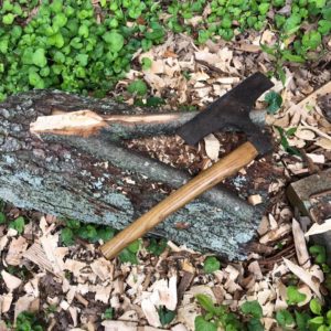 Photographed is the fork of a fallen tree limb (the fork is where two branches meet and form an open V shape), atop the fork rests a hewing axe which is a wooden handled axe whose blade is rectangular in shape. These items have been photographed atop other tree limbs and a bed of wooded grass.