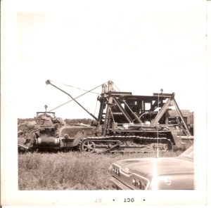 A black and white polaroid photo dated OCT '67. The photo shows a ditch digging machine used to help lay pipeline, and the rear end of a classic car in a grassy fielded area.