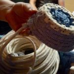 Photographed is a coiled rope basket being made. The basket's bottom is blue while the material on the side is a white and blue striped pattern. Holding this smaller coiled basket is Robin Reichardt's hand, below the basket is the white rope which the patterned basket is being made from.