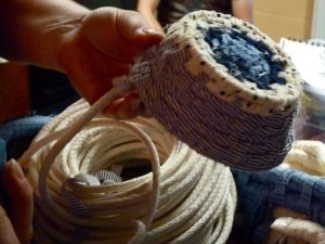 Photographed is a coiled rope basket being made. The basket's bottom is blue while the material on the side is a white and blue striped pattern. Holding this smaller coiled basket is Robin Reichardt's hand, below the basket is the white rope which the patterned basket is being made from.