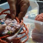 A photograph of coiled twine being made into a basket. Photographed are Robin Reichardt's hands holding orange coiled twine folded around pink cloth with colorful characters on it. Behind her hands are two balls of yarn in a clear storage bin.