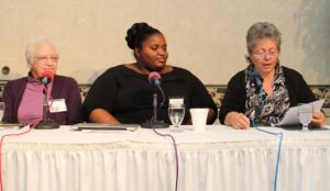 Panel photograph from the 2015 Missouri Folklore Society meeting in Jefferson CIty, MO. From left to right sits Mary Peura, Angela J. Williams, and Sarah Denton. Mary Peura is an elderly white woman with white hair, she wears thin metal framed glasses and a magenta turtleneck sweater beneath a plum colored cardigan. Beside her Angela J. Williams sits, she wears a short sleeve black top and has her dark black hair breaded in an updo on her head. Beside her Sarah Denton sits reading from a piece of paper. Sarah Denton is a middle aged white woman with salt n pepper hair, she wears thin metal framed glasses and a black and white patterned shirt tucked under a black cardigan. In front of each panelist sits glasses of water and microphones.