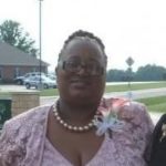 Image of Evelyn Pulliam; from the chest up. Evelyn is an older Black woman, she wears a light pink lace patterned top and matching over jacket. She wears a thick pearl necklace, and has a white broach on her shirt. She wears thick plastic framed glasses, hair is tied back in braids.