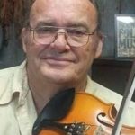 Headshot photo of Gary Ray Johnson for his tribute. Gary is an elderly white male wearing silver aviator style eyeglasses. He wears a light green colored button down shirt and in front of his neck/chest area rests a wooden violin.