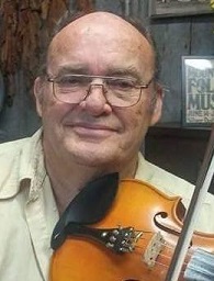 Headshot photo of Gary Ray Johnson for his tribute. Gary is an elderly white male wearing silver aviator style eyeglasses. He wears a light green colored button down shirt and in front of his neck/chest area rests a wooden violin.