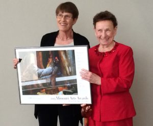 Nola Ruth and Jennie Cummings holding Missouri Arts Award poster, 2015. Nola Ruth stands left, she is an elderly white woman with short cut brown hair. She wears a black matching suit, unbuttoned to show a white shirt beneath it. She wears thin wire glasses and is smiling. Beside her stands Jennie Cummings, in a red matching suit with red beading around the collar. Jennie is an elderly white woman with brown hair tied back. She smiles, holding the other side of the framed Missouri Arts Awards poster.