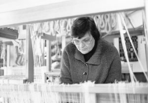 Black and white photo of Barbara Overby. Barbara is a white woman with short cut, face framing dark hair. She wears wide clear framed glasses and a black turtleneck shirt under a brown wool coat with gold buttons. She is looking down at the white yarn which hangs from a loom, which appear at the forefront of the photo.
