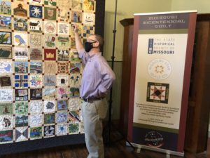Photo from the Missouri Bicentennial Quilt exhibition. Standing in front of a patchwork quilt is a middle-aged white man, he wears a longsleeved lilac button down shirt tucked into khaki pants, a dark brown leather belt at his waist. He wears a black face mask and thin wired glasses. He faces left, one arm leaning against the patchwork quilt. The patchwork quilt features iconic elements of Missouri such as the state bird, various map outlines, and churches.