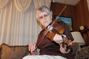 Vesta Johnson is photographed playing fiddle at a home session during a Traditional Arts Apprenticeship Program site visit. Vesta is an elderly white woman with white hair, peppered with black at the root. She has a focused expression fixed on her fiddle, wearing a pair of thin metal framed glasses, a plain dark brown tee shirt, and a pair of white pants. Vesta sits in the foreground of the photo on a brown metal chair, behind her is an array of home decor (couch and pillows, white curtains, and a wood paneled wall with a landscape painting).