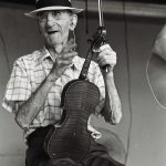 Photographed in black and white, Albert Spray sits with a fiddle resting in his lap. Albert Spray is an elderly white man, he wears a white fedora with a black band around the base. A white button down with a plaid pattern, tucked into a pair of lighter colored slacks. He holds a bow in one hand, the other gesturing to someone off camera.