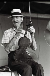 Photographed in black and white, Albert Spray sits with a fiddle resting in his lap. Albert Spray is an elderly white man, he wears a white fedora with a black band around the base. A white button down with a plaid pattern, tucked into a pair of lighter colored slacks. He holds a bow in one hand, the other gesturing to someone off camera.
