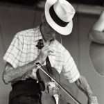 Photographed in black and white, Albert Spray is an elderly white male. He wears a white fedora with a black band around its base, a white short sleeve button down shirt with a checkered pattern, tucked into a pair of slacks. Against his lap rests a fiddle, which he is playing.