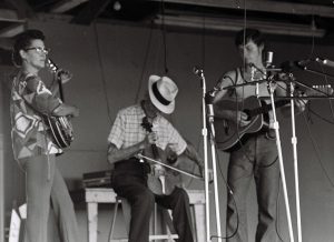 Photographed in black and white during the Braymer Fiddle Convention from left to right: Lena Hughes, Albert Spray, and an unidentified guitarist. Lena Hughes (left) is a middle aged white woman with dark brown hair tucked back in a bun, she wears a geometric patterned short sleeved top with a pair of light colored slacks. In Lena's hands is a banjo. Beside her, Albert Spray sits playing the fiddle. He is an elderly white man wearing a white fedora with a black band around the base, a white short sleeve patterned shirt, tucked into light colored slacks. Beside him stands an unnamed guitarist, he is a white teen male wearing a white tank top with black outlining the sleeves and neckline tucked into a pair of jeans. In his hands is an acoustic guitar. An array of microphones on stands sit in the foreground of this photo.