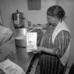 Photographed in black and white, Lillie Mabel Hall is photographed cooking for a church's Christmas dinner in 1992. Lillie Mabel Hall is an elderly Black woman, she wears a black dress with white stripes forming a checkered pattern. She wears hoop earrings and a hairnet, a white sleeveless vest completes her look. She holds a bag of active dry yeast in one hand and is adding ingredients to a large mixing bowl with the other hand.