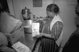 Photographed in black and white, Lillie Mabel Hall is photographed cooking for a church's Christmas dinner in 1992. Lillie Mabel Hall is an elderly Black woman, she wears a black dress with white stripes forming a checkered pattern. She wears hoop earrings and a hairnet, a white sleeveless vest completes her look. She holds a bag of active dry yeast in one hand and is adding ingredients to a large mixing bowl with the other hand.