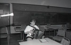 Black and white photo from a McNally Apprenticeship Program site visit. Photographed witting at a table, is McNally's apprentice Margaret Gardiner. Margaret is a younger white teen, with ginger hair pulled into a ponytail - she wears bangs in the front. In her hands is an Irish button box accordion, she pushes its buttons while staring off screen.