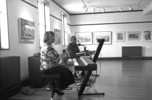 Photographed in black and white, apprentice Shannon White and master artist Charles Williams sit behind electric keyboards during a performance. Left, Shannon White appears: She is a middle aged Black woman with straight black hair that barely meets her shoulders. She wears a patterned long sleeved shirt, black flowy pants, and black high heels. She sits behind a piano with folded arms. Right, and in the background from Shannon sits Charles Williams: A middle aged Black man with salt and pepper hair. He wears a long sleeved dark v-neck style shirt, and light colored pants with brown shoes. He sits behind an electric keyboard.