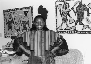Photographed in black and white, Dee Cambell Carter stands in front of African Art pieces for scale. Dee Campbell Carter is a young Black woman, she appears with straight black hair to her shoulders. She wears an African printed garment made up of light and dark hues going down in stripes, and a patterned skirt as bottoms. She stands in a Superwoman pose with her fists resting on her hips, smiling.
