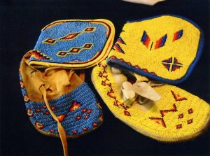 Wendi Wald's bead work photographed: a pair of Native American beaded moccasin shoes. The shoe on the left is beaded in blue, with triangular designs in yellow, orange, black, red and purple beads. The shoe on the right is a yellow beaded shoe with different patterned designs in orange, red, light blue, and dark blue beads.