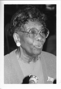 Photographed in black and white surrounded by a white border, a photo of Ann Pittman speaking into a microphone which is at her chest level. Ann Pittman is an elderly Black woman. She appears with slightly curled black hair which curls around her face. She wears large, clear framed glasses and has a large reflective earring in her ear. She wears a light colored shirt with a slightly darker sweater over it. She wears a thin chained necklace with a large pendant at the end, and a pendant which reads JESUS on the right side.
