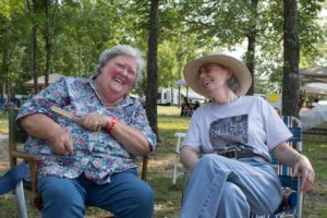 Photographed sitting beside one another outside at a festival, Marideth Sisco and Suzanne Rayfield McKenzie are laughing. Marideth (left) is an elderly plus-sized white woman with white hair and eyebrows. She wears a floral printed short-sleeve buttondown shirt with medium washed denim jeans. Suzanne is an elderly white woman with gray hair peaking out from beneath a large-brimmed white hat. She wears thin framed metal glasses, a white short-sleeved graphic tee shirt tucked into light-wash denim jeans.