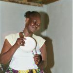 Photographed in an unfinished basement with wooden slats above her head, Mary Luka Kemir stands peering to the right smiling at someone off-camera. Mary is a taller Black woman, her dark black hair is tied in the back in a ponytail. She has high shiny cheeks, and wears a short sleeved white shirt tucked into an African patterned purple and yellow skirt. Mary wears a beaded necklace and holds a saucer to it, scraping the plate against the beads to make music.