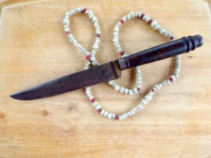 Photographed against a light wooden surface, a knife rests atop a folded beaded necklace. The knife appears with a dark stained wooden handle, it is rounded with round detailing on the end. The blade of the knife is a dark metallic gray, pointed at the end. The beads are mostly white with accents of red and dark red beads.