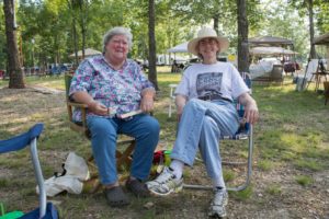 Photographed sitting beside one another outside at a festival, Marideth Sisco and Suzanne Rayfield McKenzie appear staring at the off-screen photo taker. Marideth (left) is an elderly plus-sized white woman with white hair and white eyebrows. She wears a floral printed short-sleeve buttondown shirt with medium washed denim jeans and brown crocs shoes. Suzanne is an elderly white woman with gray hair peaking out from beneath a large-brimmed white hat. She wears thin framed metal glasses, a white short-sleeved graphic tee shirt tucked into light-wash denim jeans, and running shoes with long white socks.