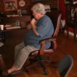 Photographed from inside, Marideth Sisco sits in a padded brown rolling chair. She faces left with her legs crossed at the ankles and hands clasped together against her chest. Marideth is an elderly plus-size white woman with white hair. She wears thin framed glasses, a blue short sleeved shirt, and a light wash brown pant, on her feet are brown loafers.
