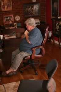 Photographed from inside a home studio, Marideth Sisco sits in a padded brown rolling chair. She faces left with her legs crossed at the ankles and hands clasped together against her chest. Marideth is an elderly plus-size white woman with white hair. She wears thin framed glasses, a blue short sleeved shirt, and a light wash brown pant, on her feet are brown loafers.