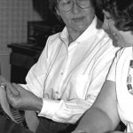 A black and white close up photo of Helen Jeffrey and her apprentice. Helen Jeffrey takes up most of the frame, she is a middle aged white woman with dark brown hair, cut short. She wears large clear framed glasses and peers over at her apprentice with a smiling expression, lips slightly pursed. Helen ears a long sleeved white button down shirt, holding in her hands weaving fabric. Her apprentice watches Helen's hands, a smile on her face. She is a younger white woman with curly hair, a short sleeved white top and a necklace made of individual rolls of thread. She holds onto fabric being weaved.