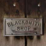 Photographed against a dark stained wooden background, a gray rectangular metal plate reads "BLACKSMITH SHOP". The plate is held against the wood with 4 metal screws, two on top and two on bottom.