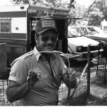 Photographed in black and white, Roger Curry sits in the foreground of the photo. Roger is a middle-aged tan skinned man, he appears wearing a cap which reads "BOSCH Spark Plugs" wearing dark aviator style glasses and a light colored short sleeve workshirt. Roger appears posing for the photo with a smile, he holds a "U" shaped basket handle in both hands. Behind him appears a pick up truck with camper shell, beside three other vehicles