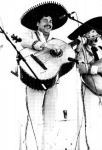 Photographed in black and white, Juan Torrez appears in mariachi styled clothing. He is a Latino man with a large black mustache and black hair, he appears smiling. He holds a large Mariachi bass guitar, one hand holding the instrument's neck, the other strumming.