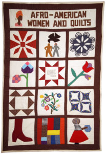 A patchwork quilt photographed in color. There is a white rectangular patch at the top of the quilt that features from left to right the outline of an Afro-American woman, the outline is faceless and only appears from the shoulders up. The caricature wears a pink top, an orange head-wrap, and gold hoop earrings. The rectangular section reads from left to right, top to bottom: Afro-American Women and Quilts. Following this rectangle, the rest of the quilt is made up of 12 square patches, aligned in 4 rows of 3 squares. The first row from left to right features a square made up of a red and white flower, a square with two woman figures and a bird flying overhead, and a blue and white patchwork flower. The second features a pink, blue, and purple flower with a long green stim, a center square with wording and a red geometric shape outlining it, and lastly a red and purple flowered patch with a long green stem. The third row features brown geometric shapes over a white background, a center square made up of red, blue, yellow, and purple flowers, and a third square featuring blue and white geometric shapes. The last row features a square with a red boot, a square made up of orange, blue, yellow, gray, and plaid rectangles forming a pattern, and the last is a silhouette of a Black woman who wears black a plaid head wrap, plaid shirt, red skirt, and black shoes. The quilt is bordered in brown.