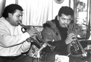 Photographed in black and white, from left to right appears Beto Lopez and his apprentice Antonio Sierra, Jr. Beto is a middle-aged Latino man, he has curly black hair which falls over his forehead, and wears a light sweater with darker pants. Beto holds a trumpet to his mouth, eyes closed in concentration as he blows into the instrument. Beside him on the couch is his apprentice Antionio, a younger Latino man with a long black mullet. He is looking up from his own trumpet to something right of the camera.