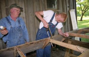 Photographed from left to right, Don Foerster is an older white male who wears a camouflage green baseball cap, a long-sleeved denim buttondown shirt, and a pair of denim overalls. Foerster wears a pair of black framed glasses and is gazing at his apprentice, Nathan Dazey's work with a slightly parted mouth. Nathan Dazey appears beside Don Foerster, a younger white male with ginger hair. He wears a short sleeved white shirt and a pair of demin overalls. In Nathan's left hand is a smaller metal handsaw. In front of the pair is the wooden structure of a jonboat. Behind the pair is a weathered wooden building.