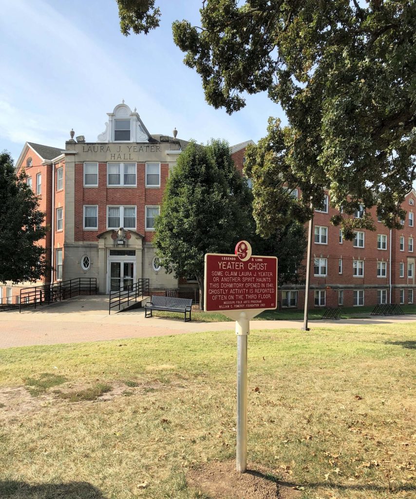 Photographed in daylight, the Yeater Ghost Legends and Lore Marker stands in the foreground of the photo. The sign itself is a golden yellow bordered red sign, the golden yellow font too small to read. Behind the sign is a the Laura J. Yeater Hall building, a large red brick and limestone building with 4 levels.