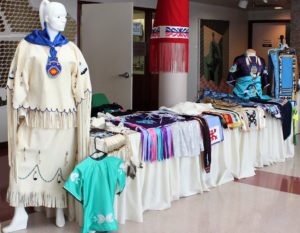 McKinney traditional clothing on display. From left to right there appears a white painted female mannequin. The mannequin wears a long sleeved white garment with royal blue trim, the sleeves and him of the garment contain long white tassled fabric. The mannequin wears a long white skirt of the same white and royal blue, Native American beadwork and white tassles fraying the skirt. Around the mannequin's neck appears a satin blue scarf, and a royal blue pendant with a red, orange, and yellow beaded design in the center. Beside the model is a table displaing other clothing items which appear in an array of white, blue, purple, pink, and red fabrics. On the far side of the table appears a headless mannequin which adorns a Navy blue and light blue garment, Native American beads hanging around the neck.