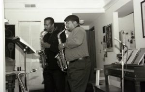 Dwayne Bosman photographed in black and white beside his apprentice. Bosman appears on the left side of the photo. Bosman is a Black male with short black hair, he wears an long-sleeve black shirt as well as long black pants. In his hands is a saxophone, which he is playing. Beside Bosman appears his apprentice, who is a lighter skinned Black male wearing a black newsboy cap. The apprentice wears a light colored sweater over a black turtleneck, dark pants with a black belt. The apprentice also holds and is playing a saxophone. In front of the pair is a black music stand, another saxophone resting on the floor beside it.