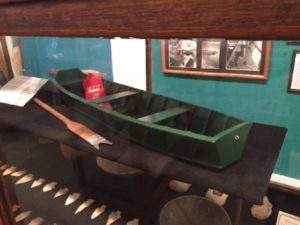 A miniature green johnboat model sits atop a black display table. The johnboat holds a red second place ribbon. On the left of the green johnboat is a brown wooden paddle. Beneath the johnboat are an array of medium sized arrow heads ranging in shape from pointy and round.